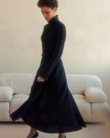 Model wearing a black ribbed cropped sweater with a long skirt, standing in a softly lit room.