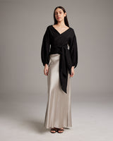 Model wearing a black peasant wrap blouse with puff sleeves and a tied waist, paired with a satin skirt.