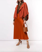 Model wearing a rust-colored bolero over a brown shirt and orange skirt, holding a small handbag.