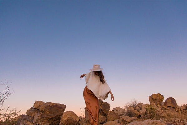 A woman in a flowing dress and hat walking among rocks at sunset