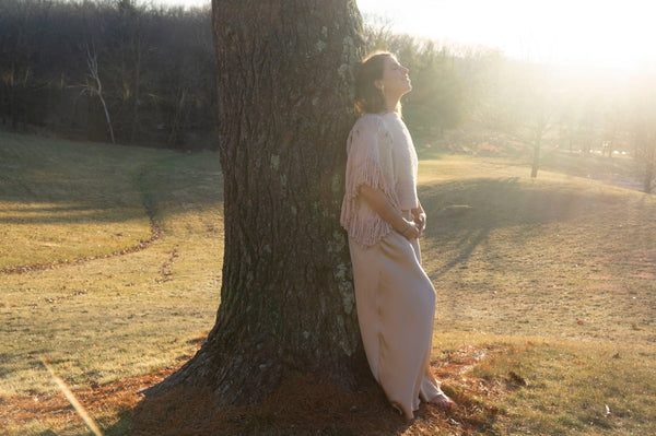 Lena Cole standing by a tree in a sunlit field, wearing a light shawl and dress