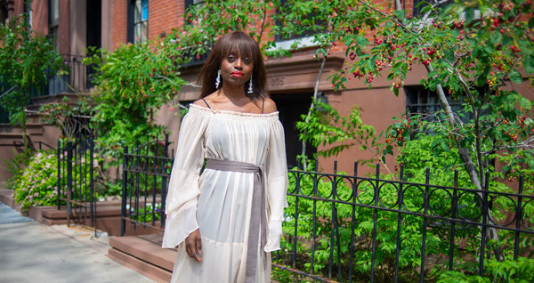 Dee Poku standing in front of a green garden, wearing a flowing white dress