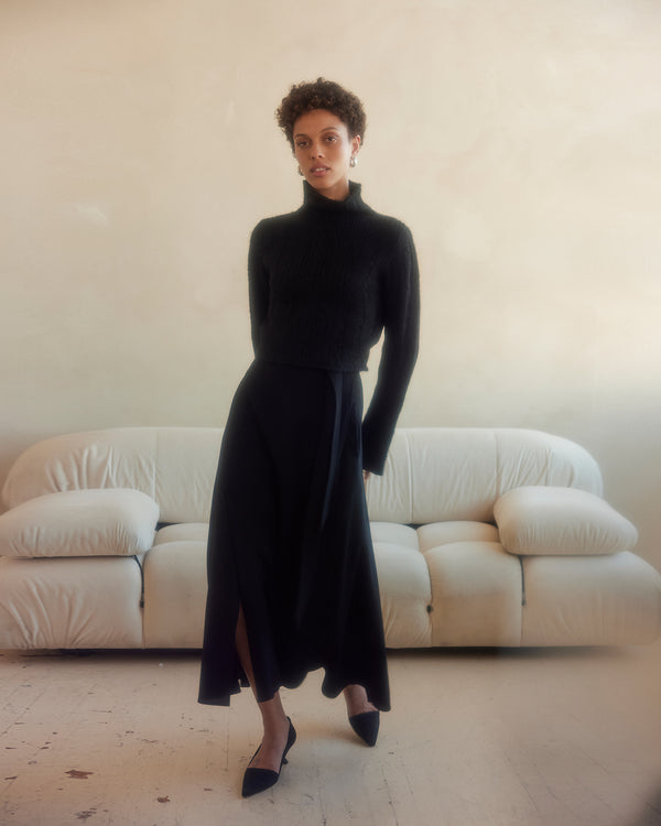 Model wearing a black ribbed cropped sweater with a black skirt, standing in a bright room.